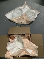 Top-down view of the open cardboard box containing ceramic plates wrapped in beige packing paper and two clear glass items placed on top outside the wrapping.