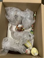 Photo showing multiple Avon ceramic dolls, a decorative vase and a mug, mostly wrapped in bubble wrap inside a box.