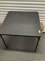 Photo of a black folding card table showing the top surface and folding legs on a carpeted floor.