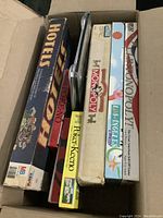 Top view of a cardboard box containing multiple boxed board games including Hotels, Pokereno, two Monopoly editions, and a set of Poker-Keeno cards.