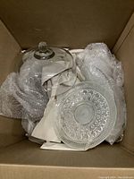 Clear glass cake trays and glass covers with decorative patterns, packed in a box with bubble wrap and paper for protection.