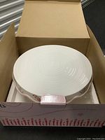 White ceramic celebration pedestal cake plate viewed from above inside original cardboard box with protective foam insert.