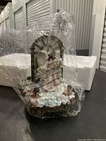 Figurine of a divine angel holding a banner, standing above cherubs and clouds, partially covered inside clear plastic packaging with visible protective styrofoam.