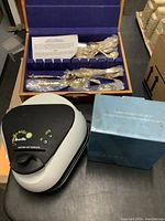 Photo showing the gold plated flatware set in wooden box with purple lining, Breville muffin maker, and garden care gift set box.