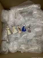 Five hand painted Victorian female figurines laid out on bubble wrap with one clear plastic wine glass among them in packaging box