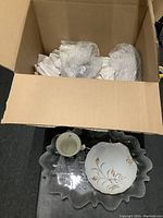 Box containing white ceramic Christmas-themed mugs and gold-rimmed plates, with a ruffled edge serving platter inside the box