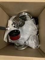 Top view of a cardboard box containing assorted pots, pans, lids, and a metal colander cup, some wrapped in bubble wrap.