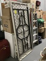 Photo of a folded large plastic folding table standing upright against a wall surrounded by boxes and other storage items. The table top is white with a textured underside and black metal folding legs.