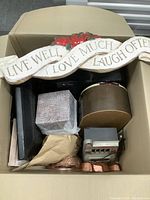 Photo shows a cardboard box containing a wooden wall decor piece with the phrase 'Live Well Love Much Laugh Often', multiple copper cake molds, a small silver vintage slot machine, a mini juke box speaker, and wrapped electronic parts.