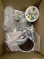 Photo showing Lladro figurine of dancing couple, tea cup, and decorative plate arranged in a box with bubble wrap