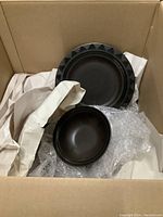 Two pieces of dinnerware - a large plate with geometric pattern at the edge and a smaller bowl, both dark colored, placed inside a cardboard box with packing material.