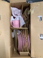 Pink dish set in cardboard packing including mugs, plates, and bowls separated by cardboard dividers.