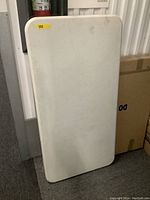 Image of a white rectangular plastic folding table with some light signs of wear, leaning against a wall in a storage area.