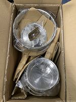 Top view of cardboard box containing multiple stainless steel pots and lids wrapped in plastic and cardboard inserts.
