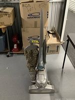 Sentria upright vacuum cleaner displayed standing upright with a patterned fabric dust bag. There are cardboard boxes and a red mixer appliance in the background.