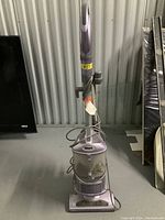 Photo of a lilac Shark upright vacuum cleaner with a clear front dustbin and power cord wrapped behind.