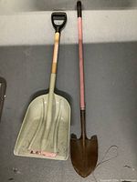 Photo showing two shovels side by side on the floor, one a plastic snow shovel and the other a metal garden shovel with wooden handle.