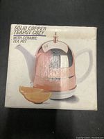 Photo of the product box showing a copper teapot cozy covering a white ceramic teapot with spout and handle, including branding and product description.