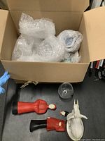 Top view of a box containing wrapped Carnival Cruise Line souvenir cups with bubble wrap, red and black limited edition tankard, small whale figurine, white ceramic elephant figurine, and clear glass cup placed on table surface below box.