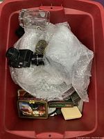 Box containing a black vintage camera, a clear glass mug, bubble-wrapped glass dishes and serving platters, and multiple decks of playing cards in tins and boxes.