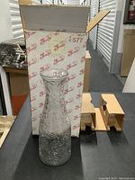 Tall clear glass wine carafe placed in front of an open vintage-style box with printed graphics.