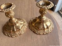 Two gold plated candlesticks, showing ornamental detail and base shape, placed on wooden surface.
