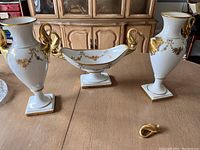 Full view of two matching porcelain vases and a dish, all decorated with gold painted floral motifs and gold swan-shaped handles. One swan handle is detached and lying in front of the dish.