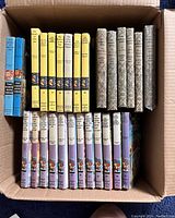Box containing 27 books arranged with hardcovers visible showing the spines of various yellow, blue, and purple books.