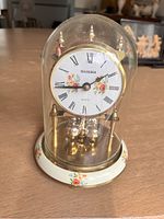 Clock under glass dome on wooden surface, showing white clock face with Roman numerals and floral pattern, brass elements and rotating pendulum balls.