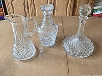 Front view of three clear crystal pieces with cut glass designs on a wooden surface: two decanters with stoppers and one pitcher with handle.