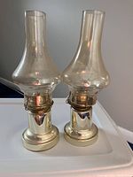 Front angled view of two matching brass oil lamps with clear glass chimneys on a white surface showcasing the brass bases and chimney shapes.