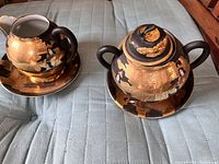 Teapot and sugar bowl or creamer on matching saucers with gold and black detailing and etching showing Chinese or Japanese styled figures and patterns.