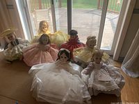 Seven full-size Madame Alexander Little Women dolls shown sitting on floor in front of window wearing various colorful and period style dresses.