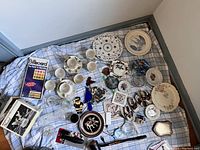 Overview of assorted porcelain plates, white teacups and saucers, small figurines, decorative plates, glass bowls, a vintage Billboard magazine and other miscellaneous items on a window sill.