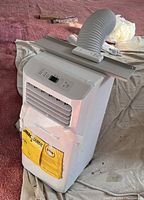 Front and angled view of white Hisense portable air conditioner with control panel, energy guide, and vent fins visible. Unit is standing on fabric drop cloth on carpeted floor.