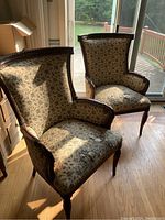 Two vintage wooden framed chairs upholstered in beige fabric with dark floral pattern, seen at an angle by a glass door.