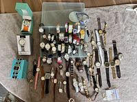 Full assortment of 64 watches displayed on fabric surface and plastic tray. Variety of band styles evident including clip, stretch, and traditional leather or metal.