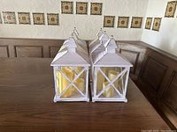 Front view of six white wooden lanterns arranged in two rows on a wooden table, showing yellow battery operated candles inside each lantern.