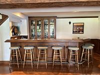 Front view of home bar with six wooden stools with padded seats and glass-door cabinetry behind the bar.