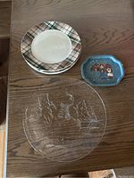 All four plates displayed on wooden table: two ceramic plaid border plates, blue rectangular plate with choir singers, clear textured glass plate with etched snowman and trees.