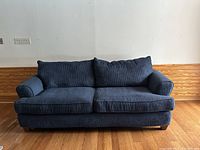 Front view of blue corduroy sofa couch showing seat and back cushions, rounded armrests, and wooden floor beneath.