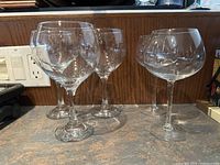 Set of 5 extra large clear wine glasses, three red wine style and two coupe style, shown on countertop.