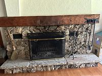Photo showing stone hearth fireplace with natural fiber broom, black metal grill screen, wall-mounted black metal fireplace tools (shovel, poker, broom) on right side.