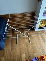 White metal folding laundry hanger with horizontal rods and X-frame base on wooden floor near shelf and sofa.