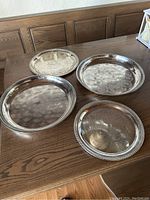 Four round metal serving trays displayed on a wooden table, showing different designs and sizes.