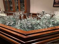 Seven rows of clear glassware on green marble and wooden table including shot glasses with logos, two low ball glasses, and small wine glasses in the background.