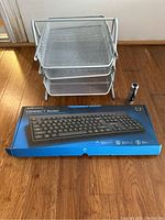 Three-tier silver metal mesh paper tray with small black flashlight and Logitech Connect Basic keyboard in box on wooden floor against white wall.