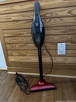 Full view of a small upright vacuum cleaner with black upper body and red base, cord coiled at the bottom, against wood panel wall.