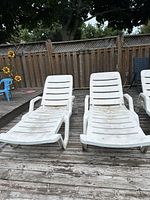Three white plastic lounge chairs arranged on a weathered wooden deck in front of a wooden fence. Chairs show dirt and wear from outdoor use.