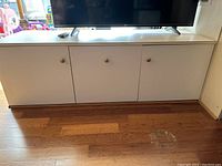 Front view of white TV stand cupboard with three doors, holding a flat-screen television and a remote control on top.